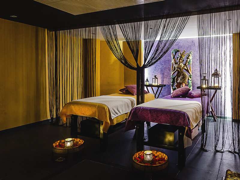 Lujosa sala de spa con dos camillas de masajes, detalles en morado y dorado, velas encendidas y una estatua.