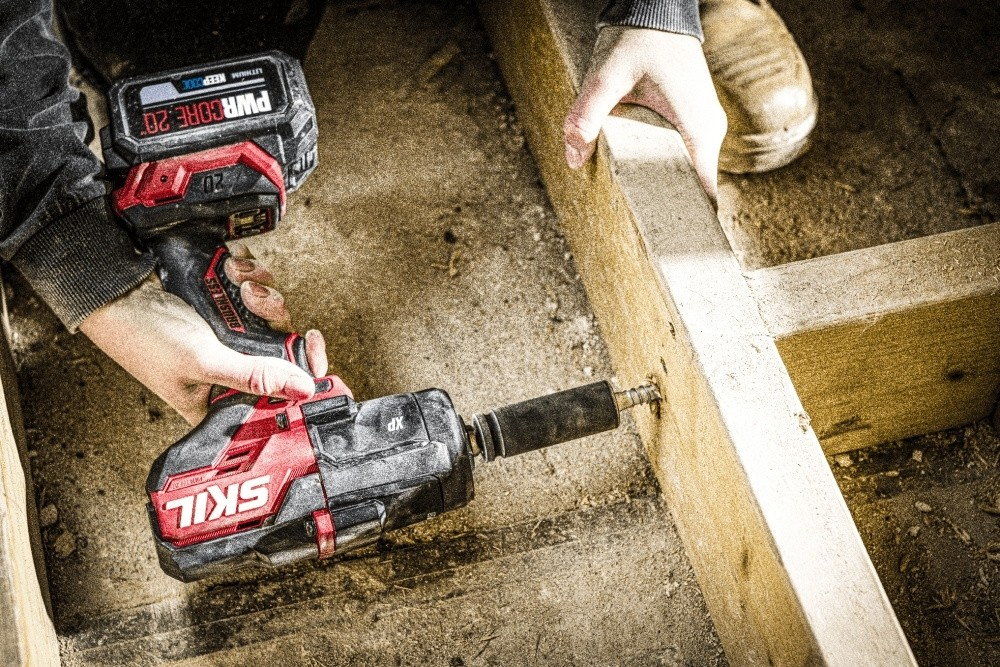 Una persona usa un atornillador de impacto inalámbrico Skil rojo y negro para atornillar un tornillo largo en una viga de madera.