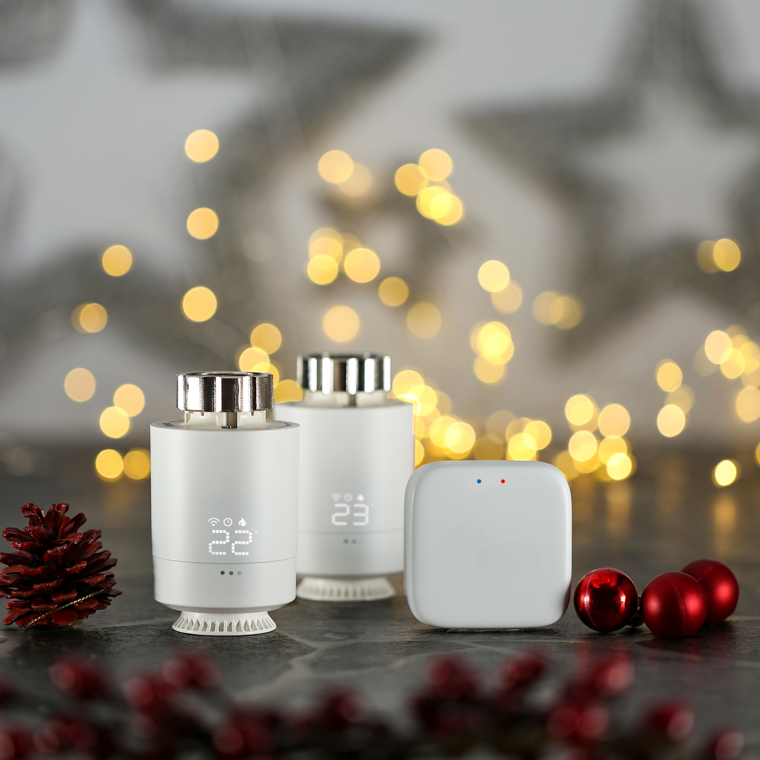 Zwei weiße Thermostate und ein Hub stehen auf einer dunklen Oberfläche vor Weihnachtsbeleuchtung.
