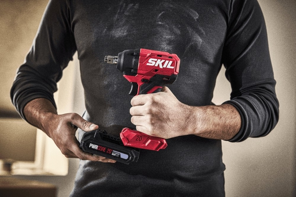 Un uomo con una maglietta nera a maniche lunghe tiene in mano un avvitatore a impulsi cordless Skil rosso e nero e una batteria.