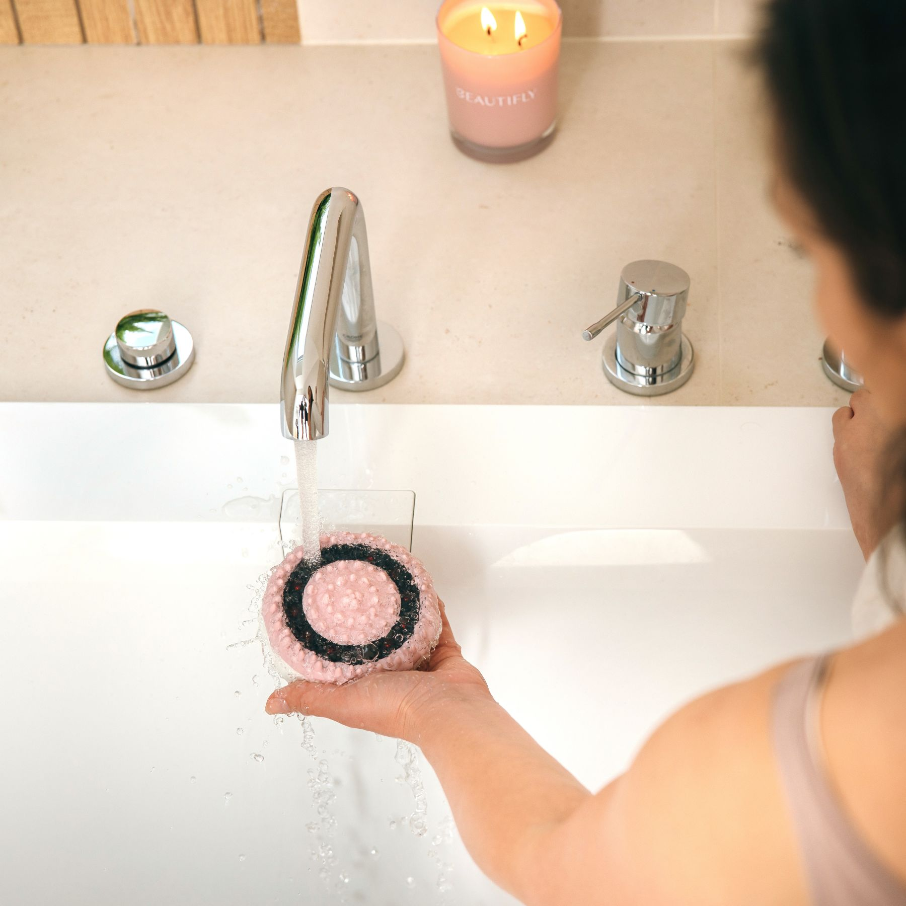 Una persona tiene una spazzola detergente rosa sotto l'acqua corrente di un rubinetto cromato, con una candela sullo sfondo.