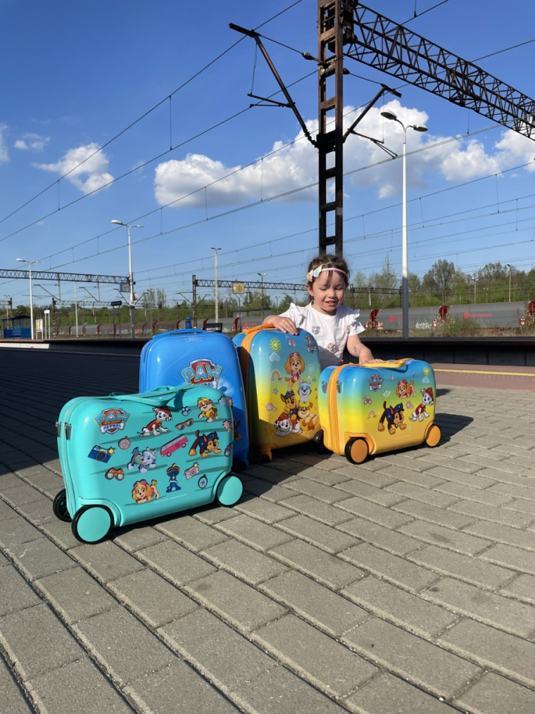 Uśmiechnięta dziewczynka pozuje na peronie z czterema kolorowymi walizkami Paw Patrol pod błękitnym niebem.