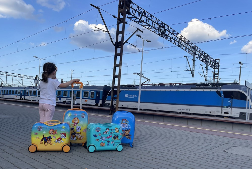 Mała dziewczynka stoi na peronie z czterema kolorowymi walizkami Paw Patrol, w tle pociąg i błękitne niebo.