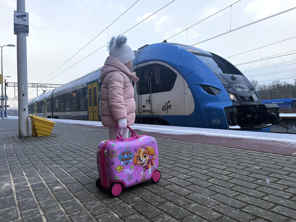 Małe dziecko w zimowych ubraniach stoi na peronie z różową walizką Paw Patrol przed niebieskim pociągiem.