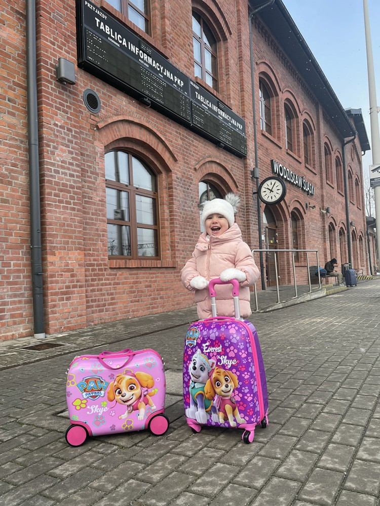 Szczęśliwa dziewczynka w różowej kurtce i czapce stoi przed ceglanym budynkiem z dwoma walizkami Paw Patrol.