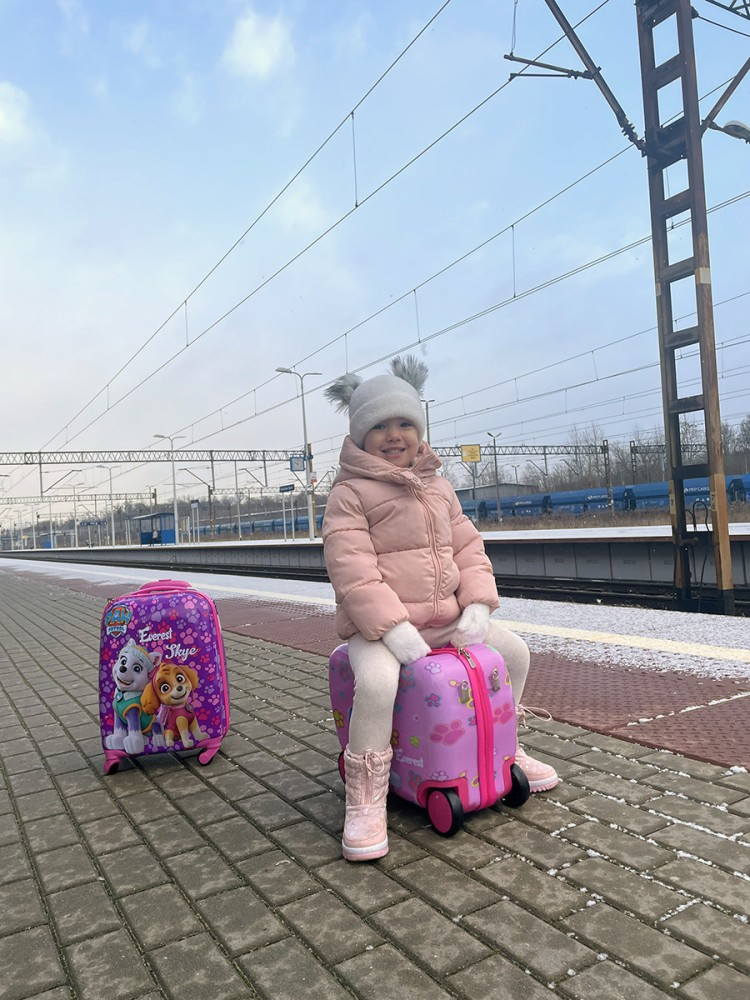 Młoda dziewczynka w różowej kurtce i czapce siedzi na różowej walizce dziecięcej na peronie.