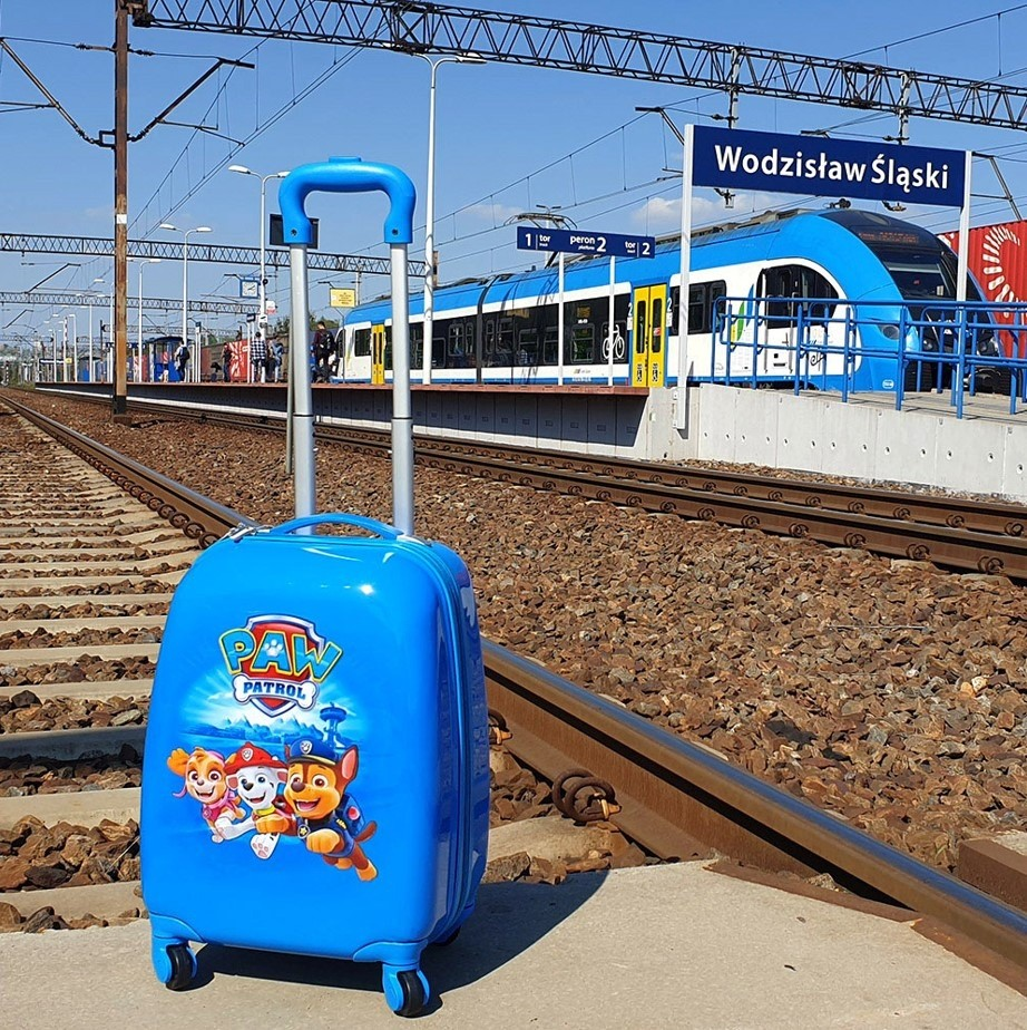 Niebieska walizka dziecieca Paw Patrol stoi na peronie kolejowym z torami i niebieskim pociagiem w tle.