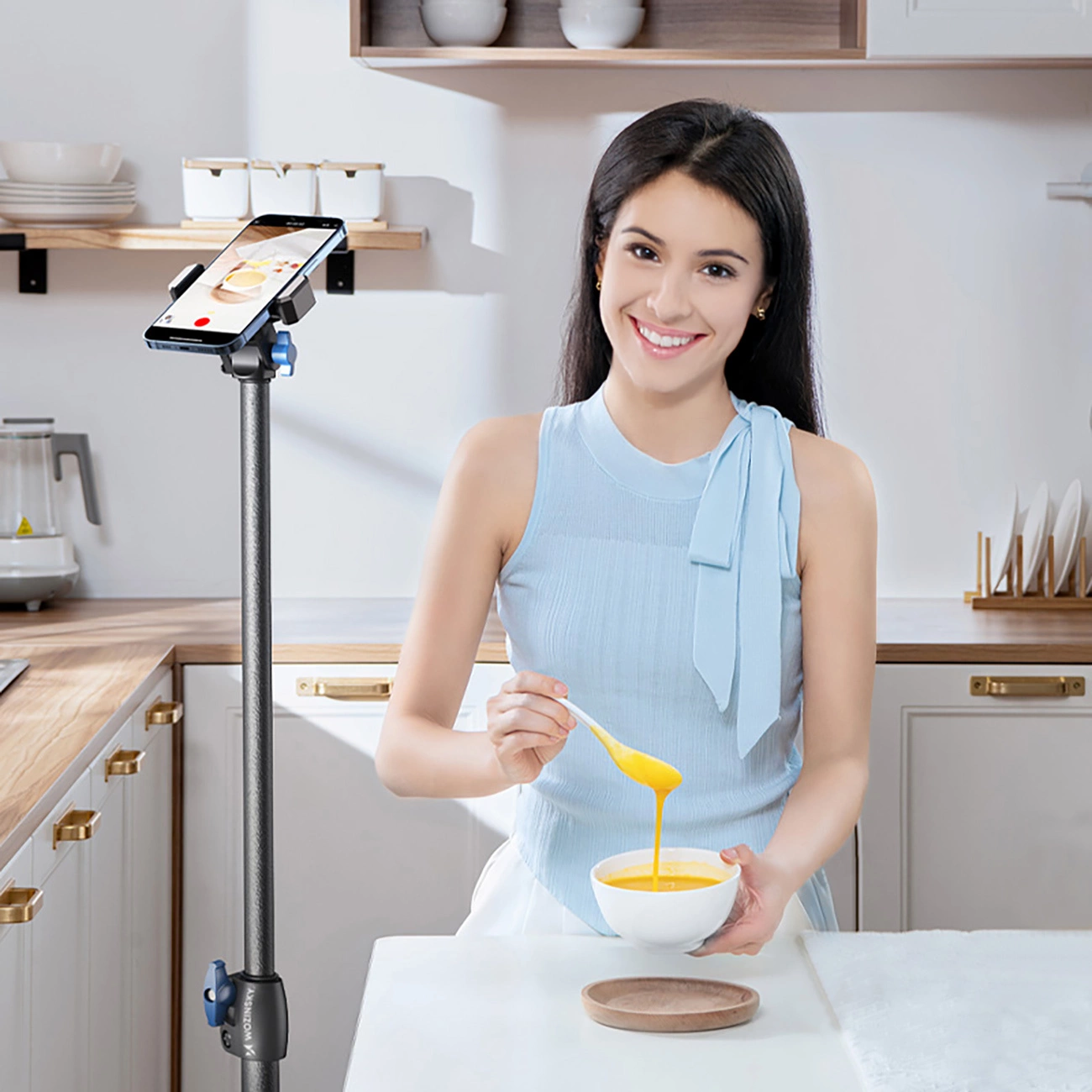 Una donna sorridente in cucina tiene una ciotola di zuppa e un cucchiaio, con uno smartphone su un supporto.