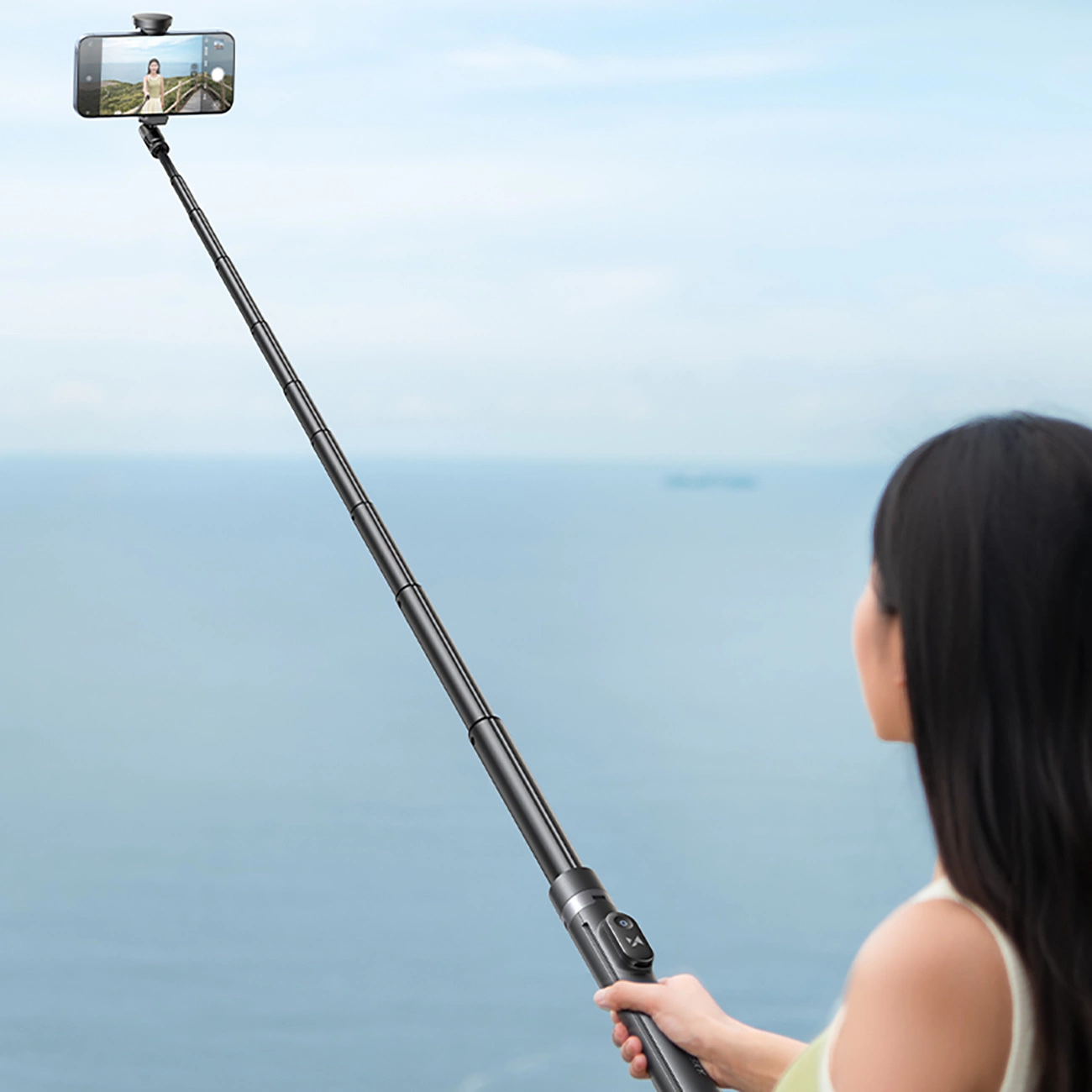 Una donna tiene in mano un bastone per selfie con uno smartphone, con il mare sullo sfondo.