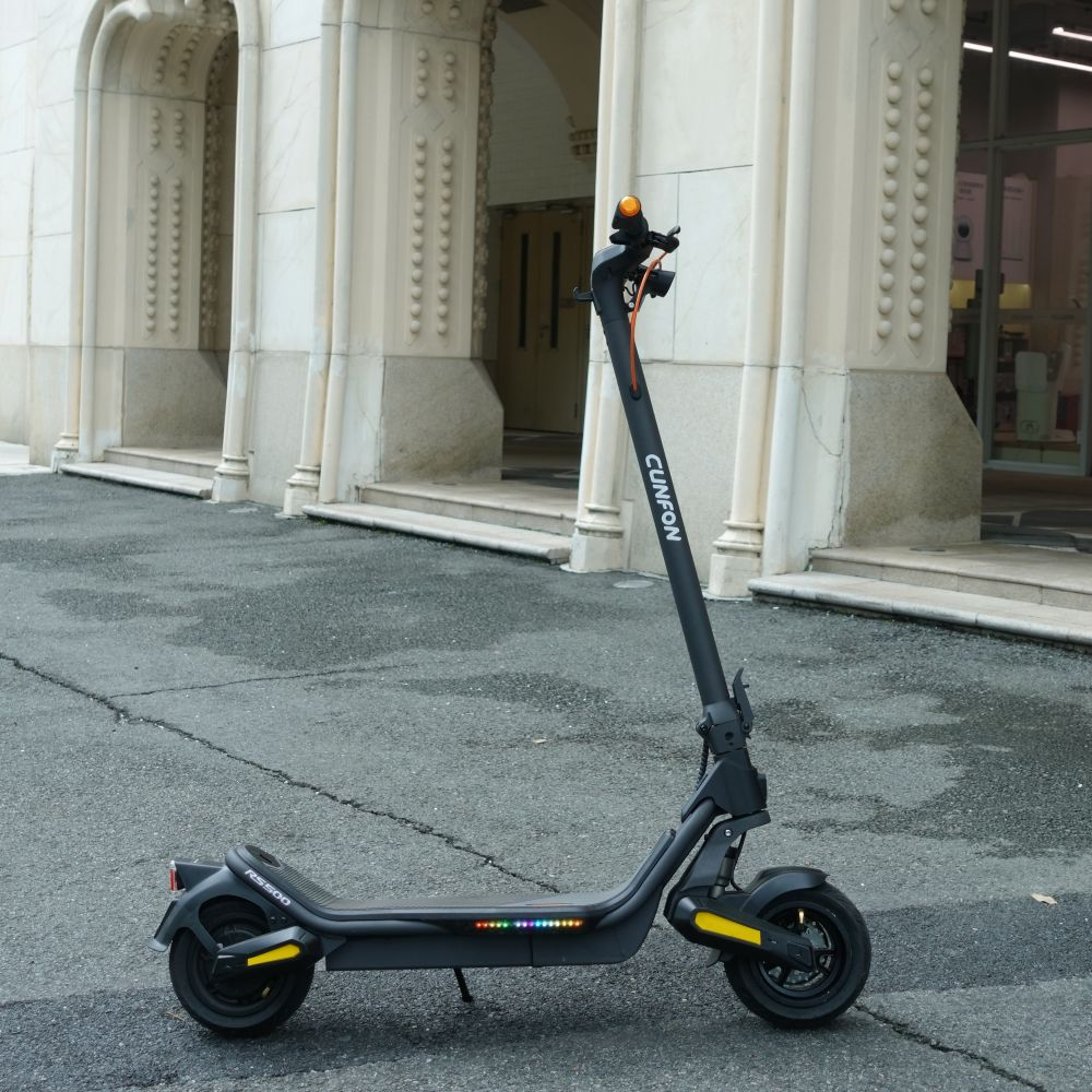 Un monopattino elettrico Cunfon grigio parcheggiato sull'asfalto davanti a un edificio chiaro con ingressi ad arco.
