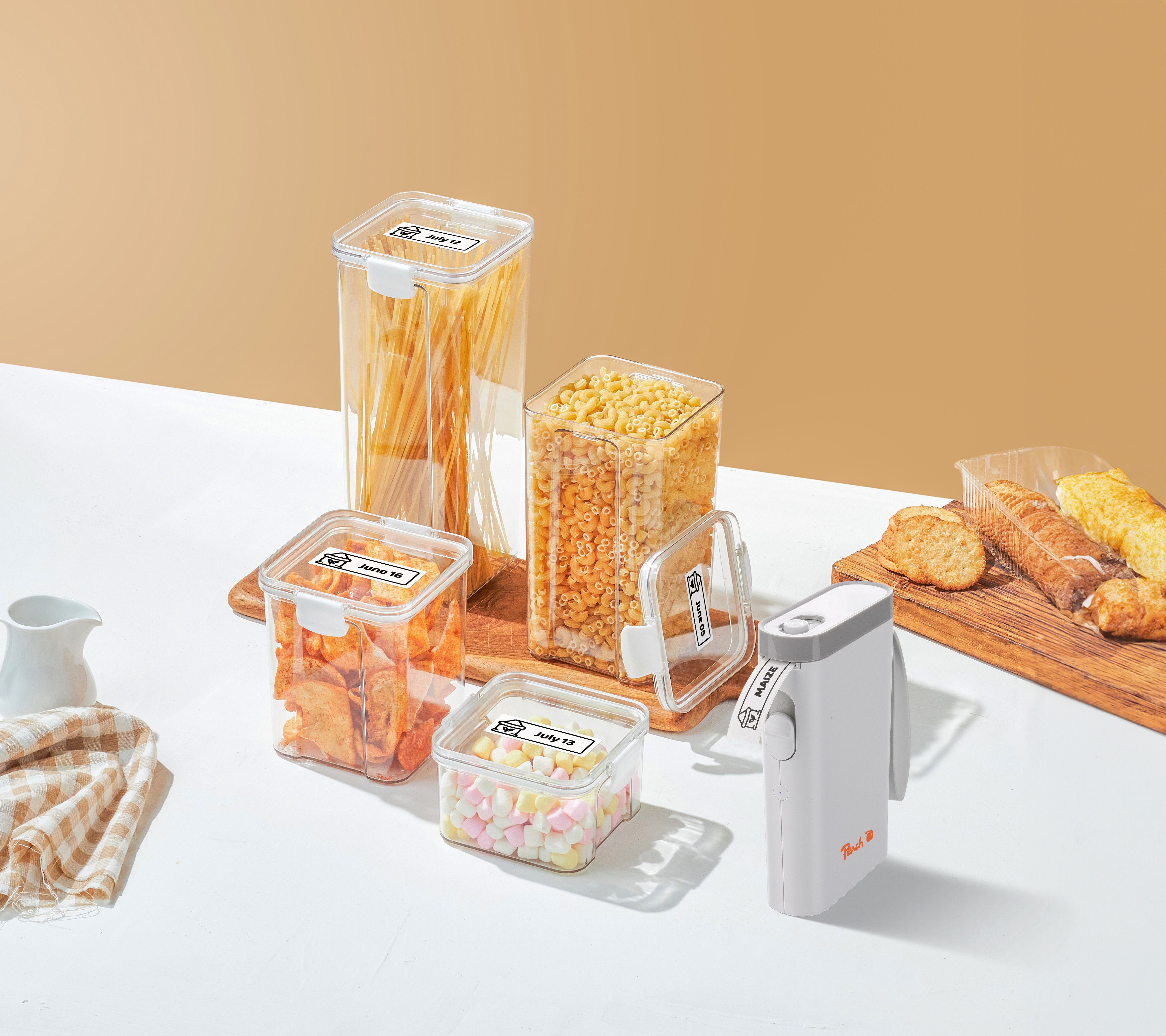 Quatre boîtes étiquetées avec pâtes et snacks, une étiqueteuse et des pâtisseries sur une table claire.