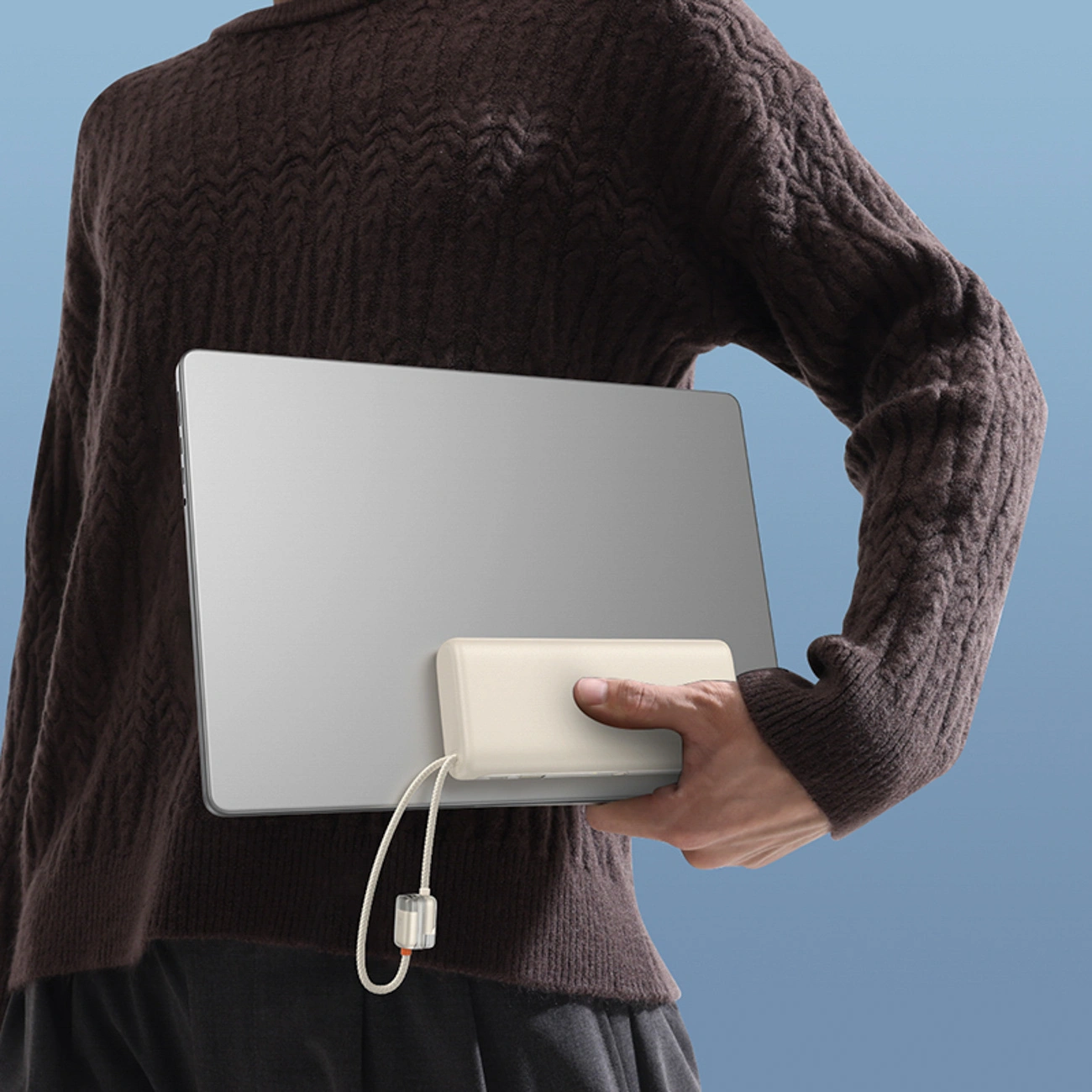 Una persona con maglione marrone tiene un laptop e un power bank beige con laccetto su sfondo blu.