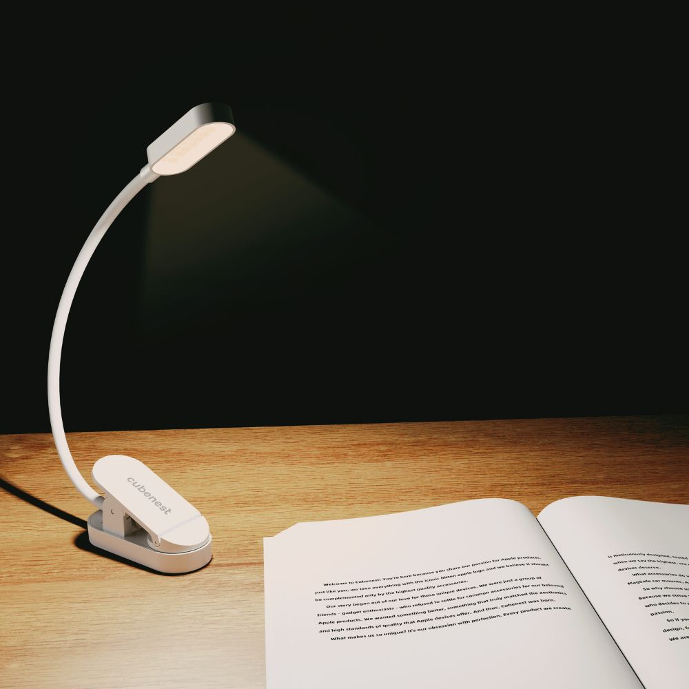Eine weiße Clip-Lampe beleuchtet ein offenes Buch auf einem Holztisch vor einem dunklen Hintergrund.