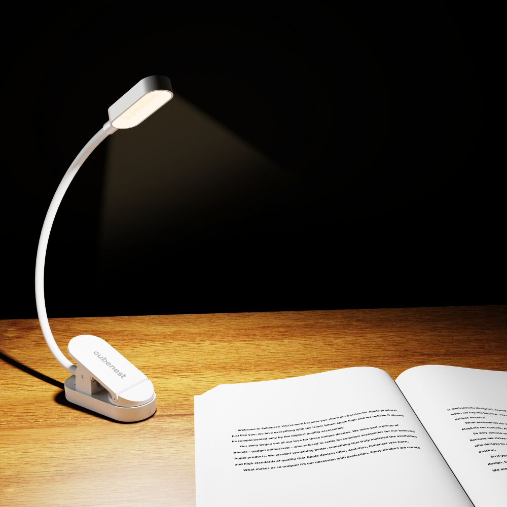 Eine weiße Clip-Lampe leuchtet auf ein offenes Buch auf einem Holztisch vor schwarzem Hintergrund.