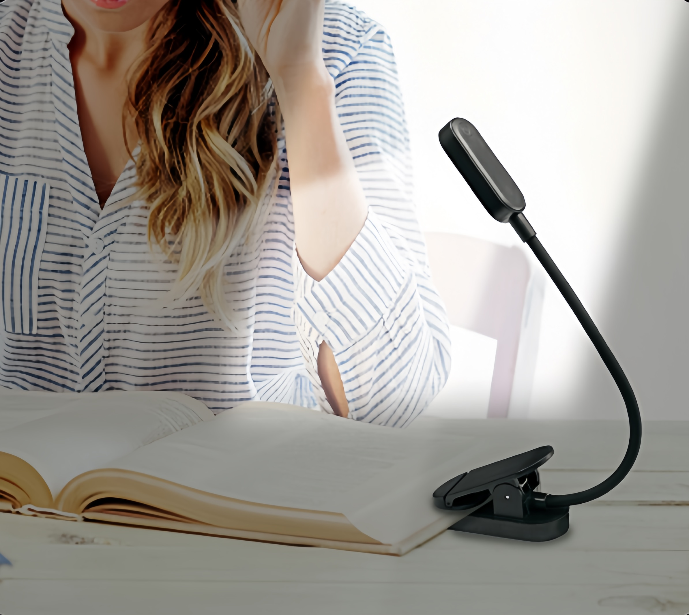Eine blonde Frau sitzt an einem Tisch mit einem offenen Buch und einer kleinen schwarzen Klemmlampe.