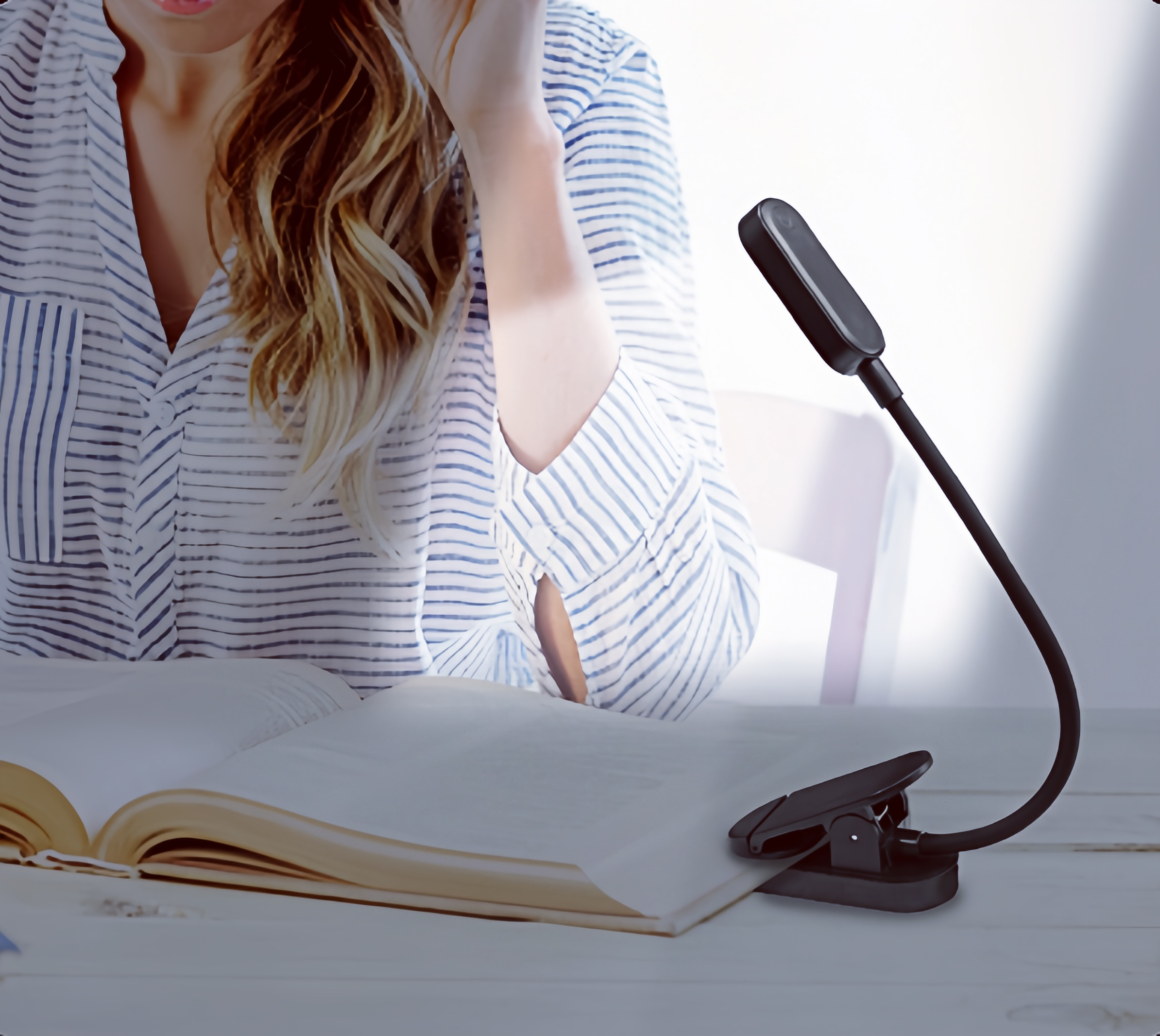 Eine Frau in blau gestreiftem Hemd liest ein Buch am Tisch, beleuchtet von einer schwarzen Klemmlampe.