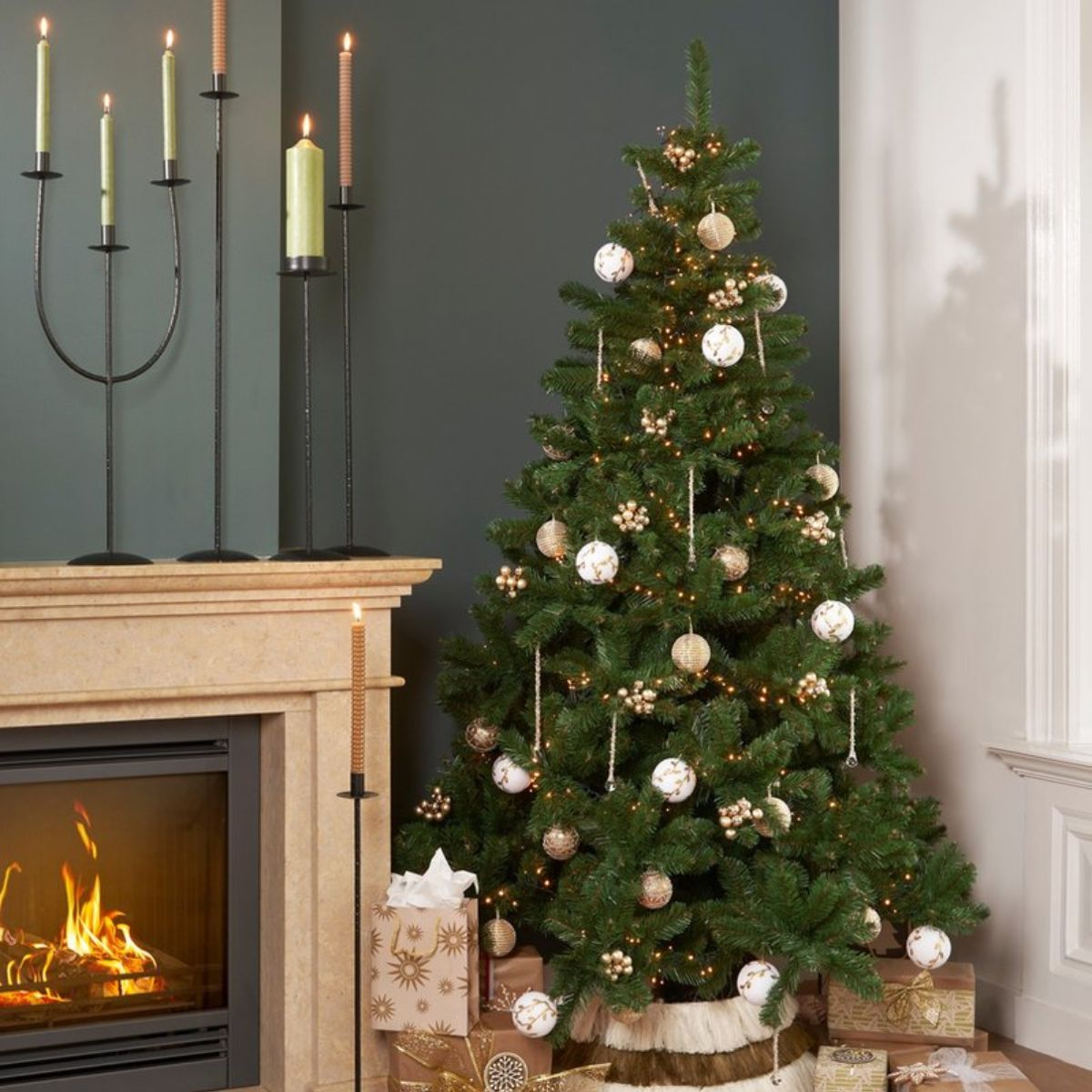 Versierde kerstboom naast een stenen open haard met brandende kaarsen, ingepakte cadeautjes en donkergroene muur.