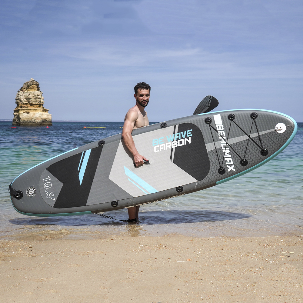 Hombre sosteniendo una tabla de SUP en la playa con rocas.