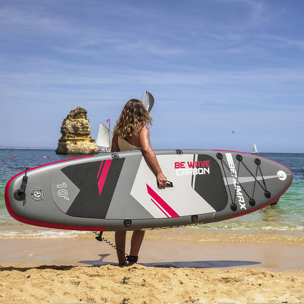 Una mujer sostiene una tabla de SUP en una playa de arena con el mar y rocas al fondo.