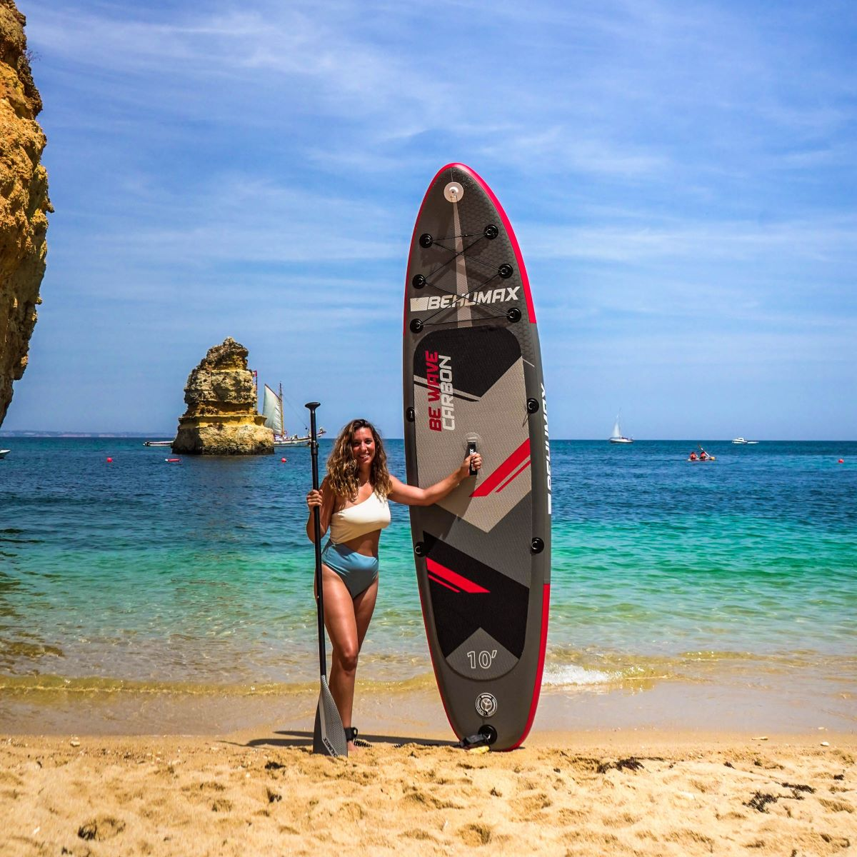 Una mujer en bikini sostiene una tabla de SUP y un remo en una playa soleada con acantilados.