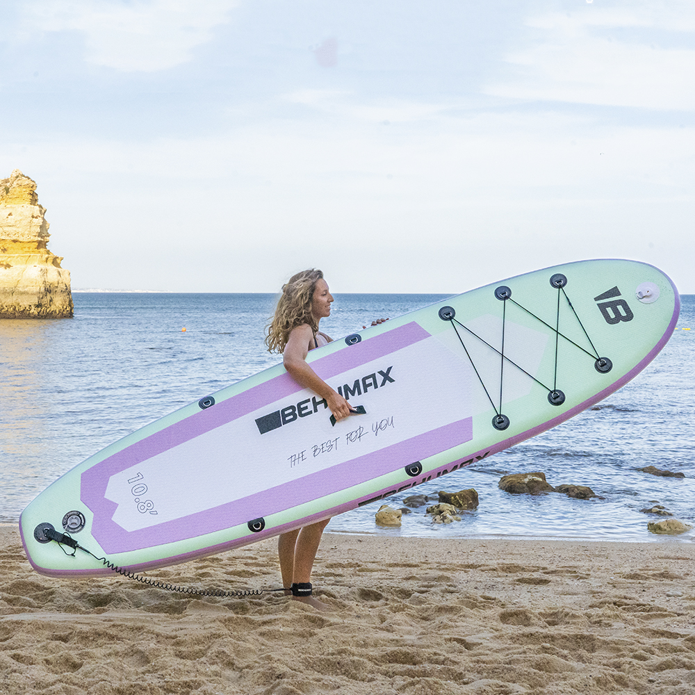 Una mujer sostiene una gran tabla SUP menta y morada en una playa de arena con el mar al fondo.