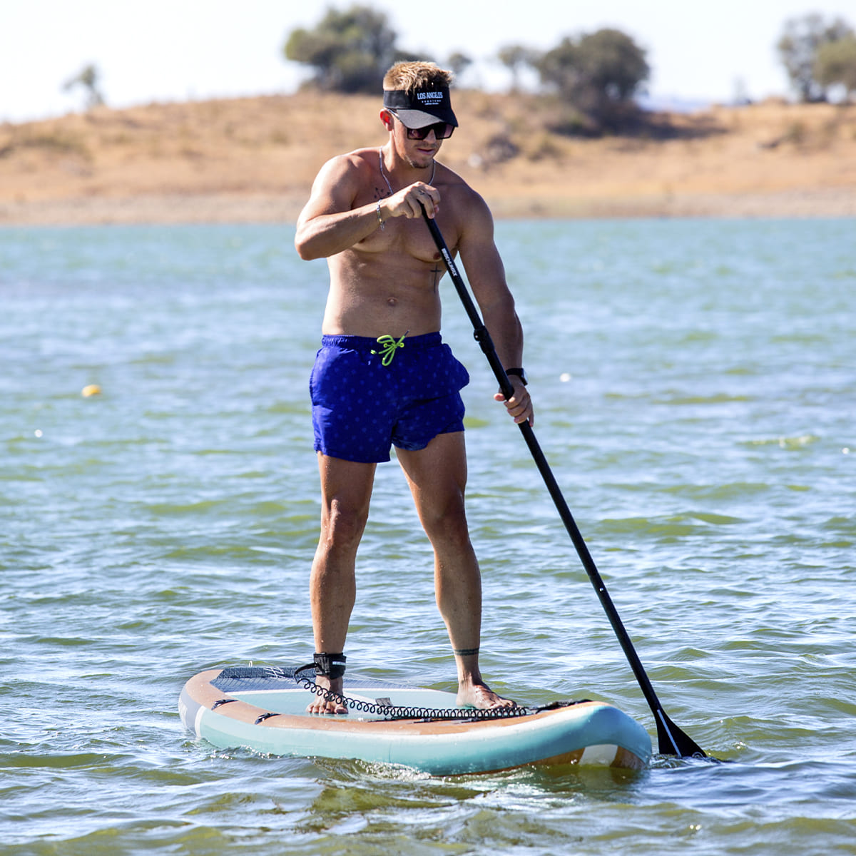 Hombre con gafas de sol y bañador azul de pie en una tabla de surf de remo en un lago.