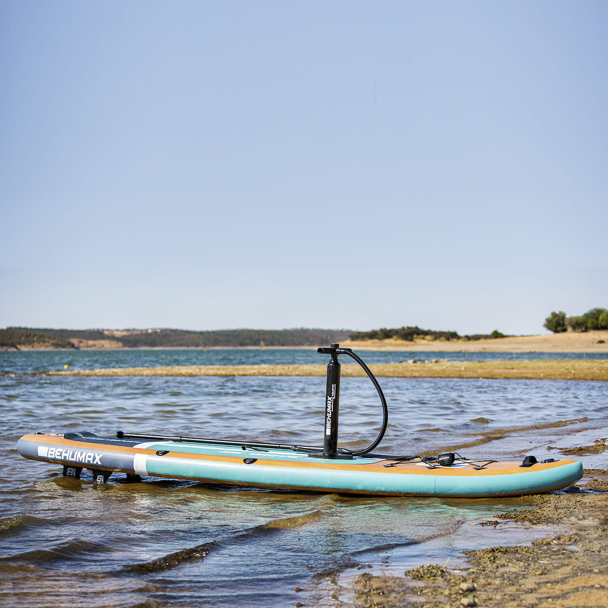 Tabla de paddle surf azul y marrón en la orilla de un lago con bomba, bajo cielo azul.