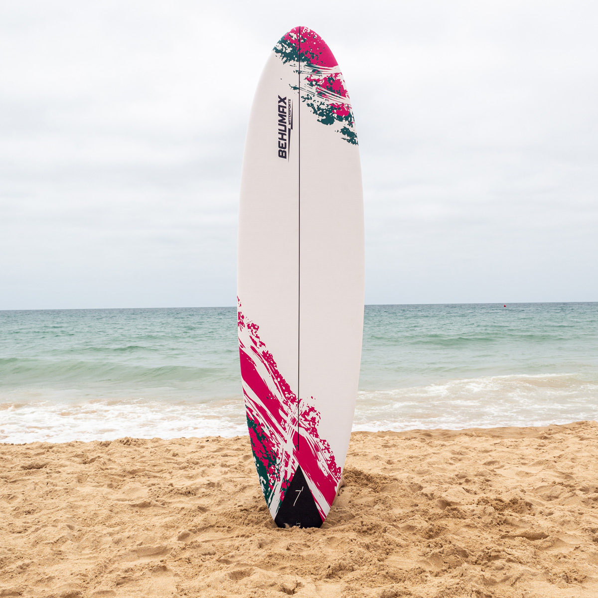 Una tabla de surf blanca con patrón rosa y turquesa está vertical en la arena de la playa con el mar al fondo.