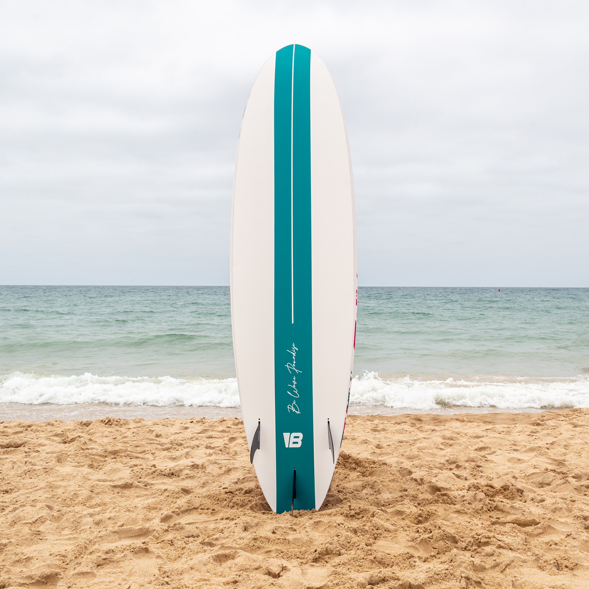 Una tabla de surf blanca con una raya verde azulada se encuentra verticalmente en la arena de una playa frente al mar.
