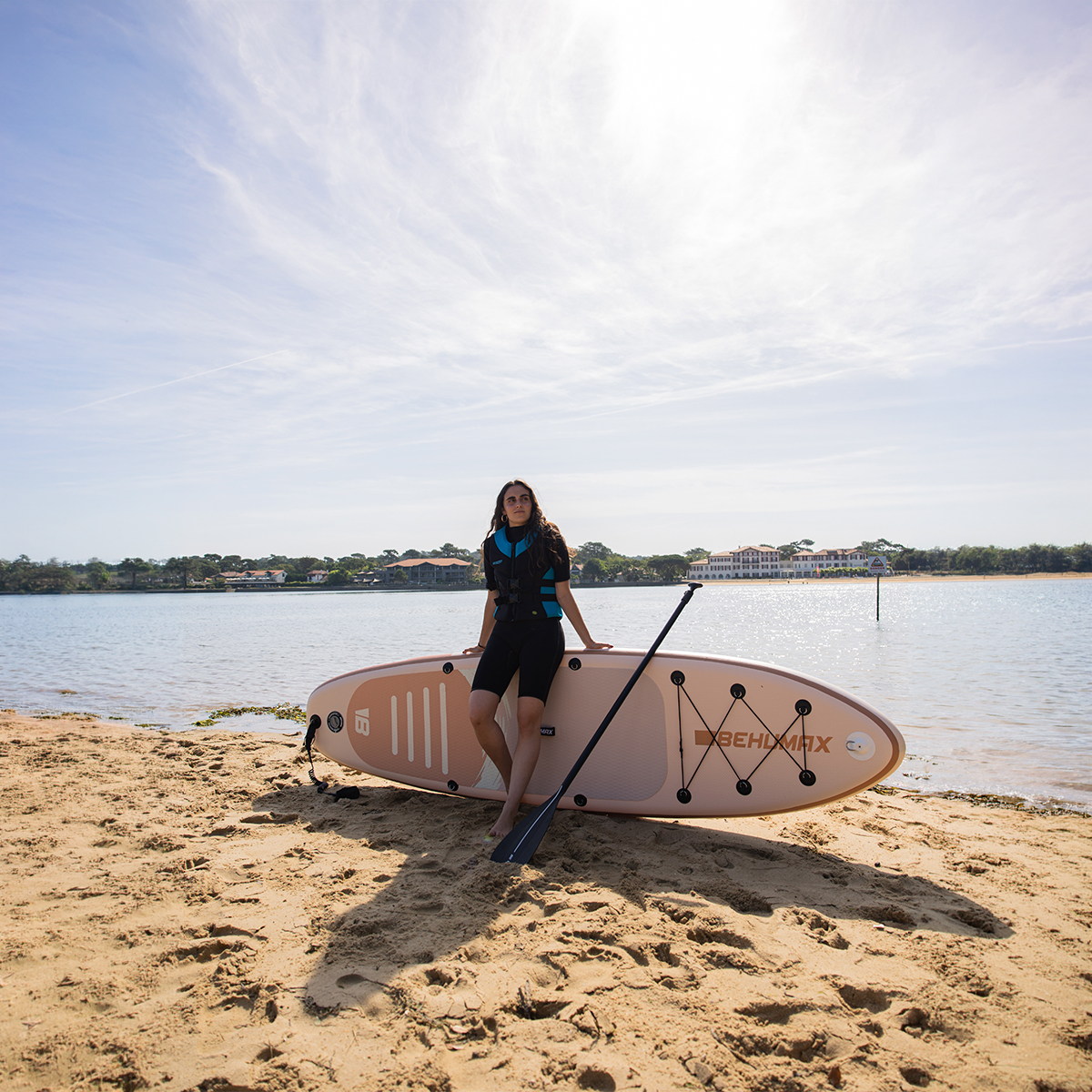 Una mujer en traje de neopreno está en una playa de arena sosteniendo un remo junto a una tabla de surf de remo rosa.