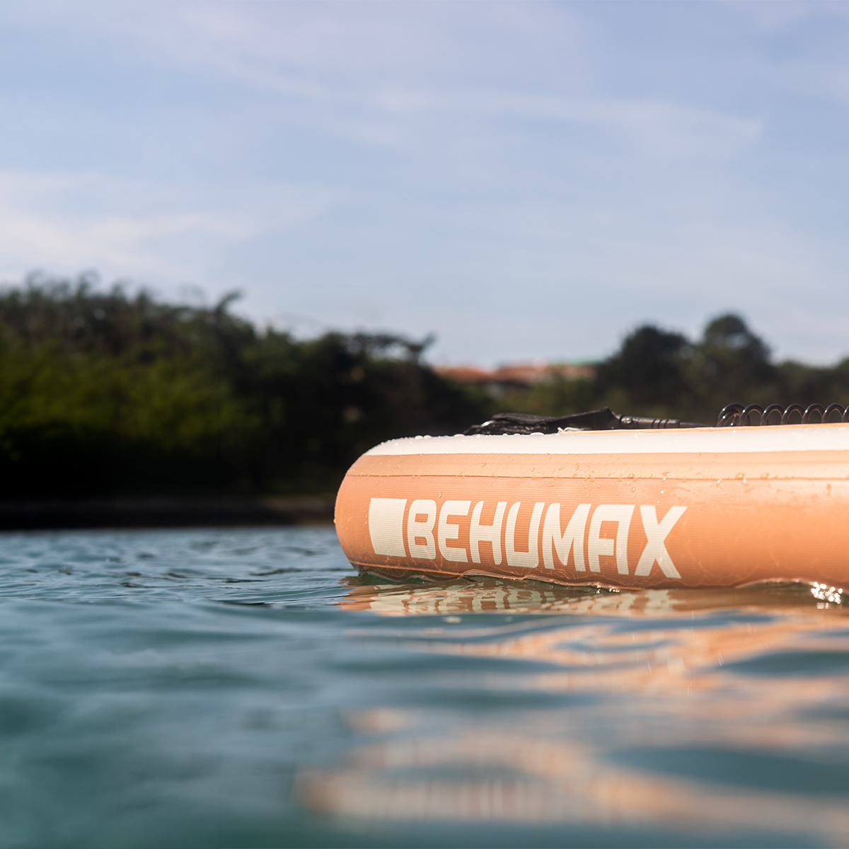Una tabla de paddle surf beige con el texto BEHUMAX flota en aguas tranquilas bajo un cielo azul, con vegetación al fondo.