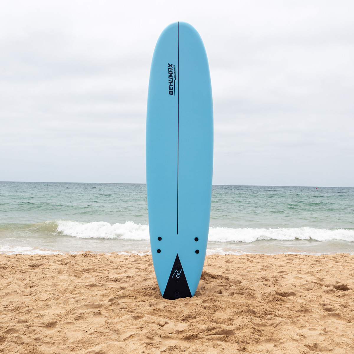 Una tabla de surf azul claro está de pie en la arena de una playa con olas tranquilas bajo un cielo nublado.