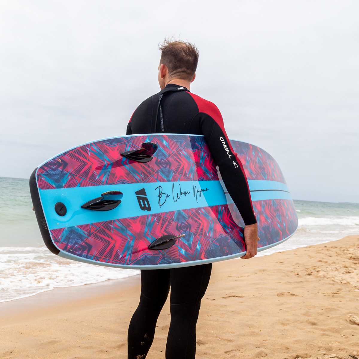 Un surfista con traje negro sostiene una tabla colorida en una playa de arena junto al mar.