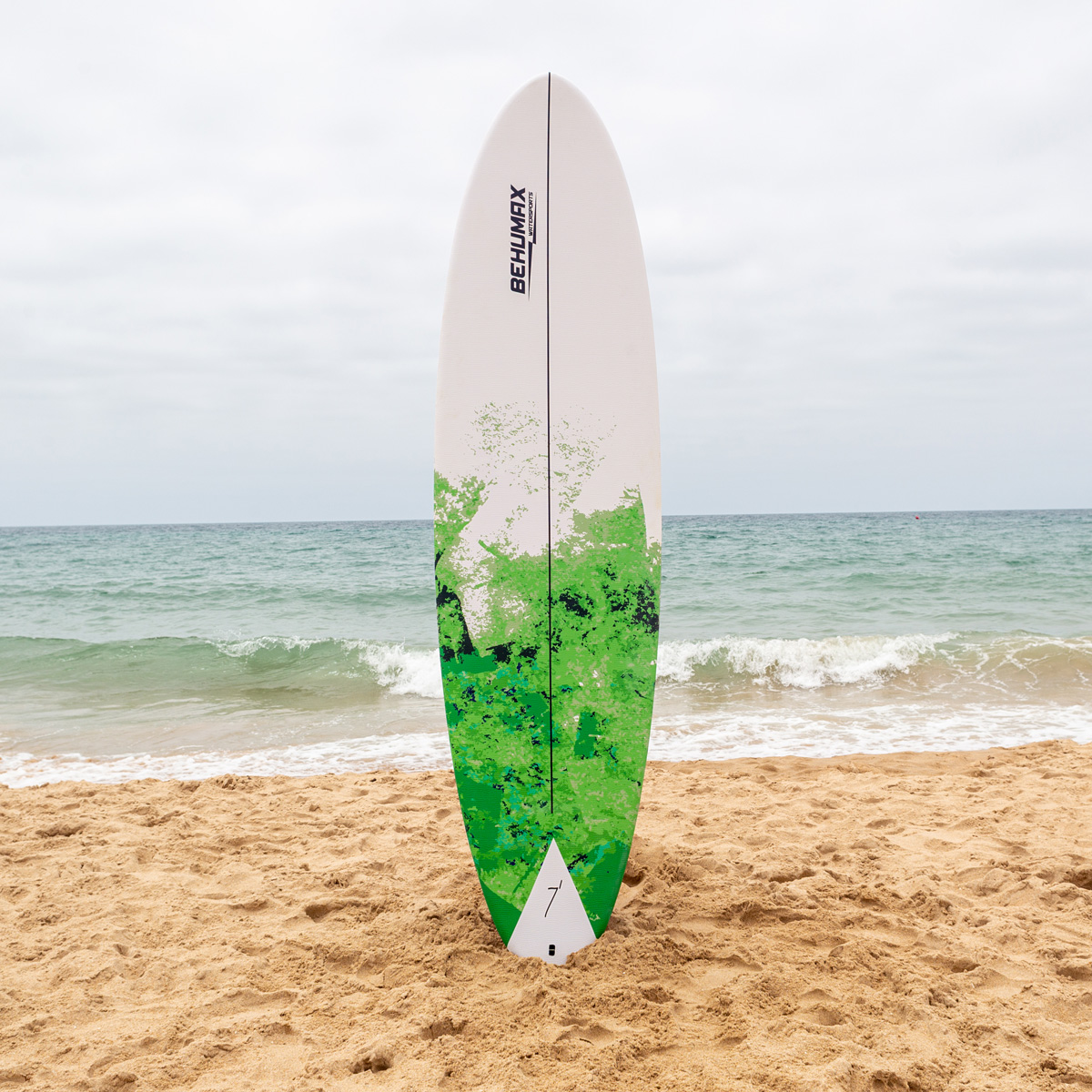 Una tabla de surf blanca y verde se encuentra en la arena de una playa con olas del mar al fondo.