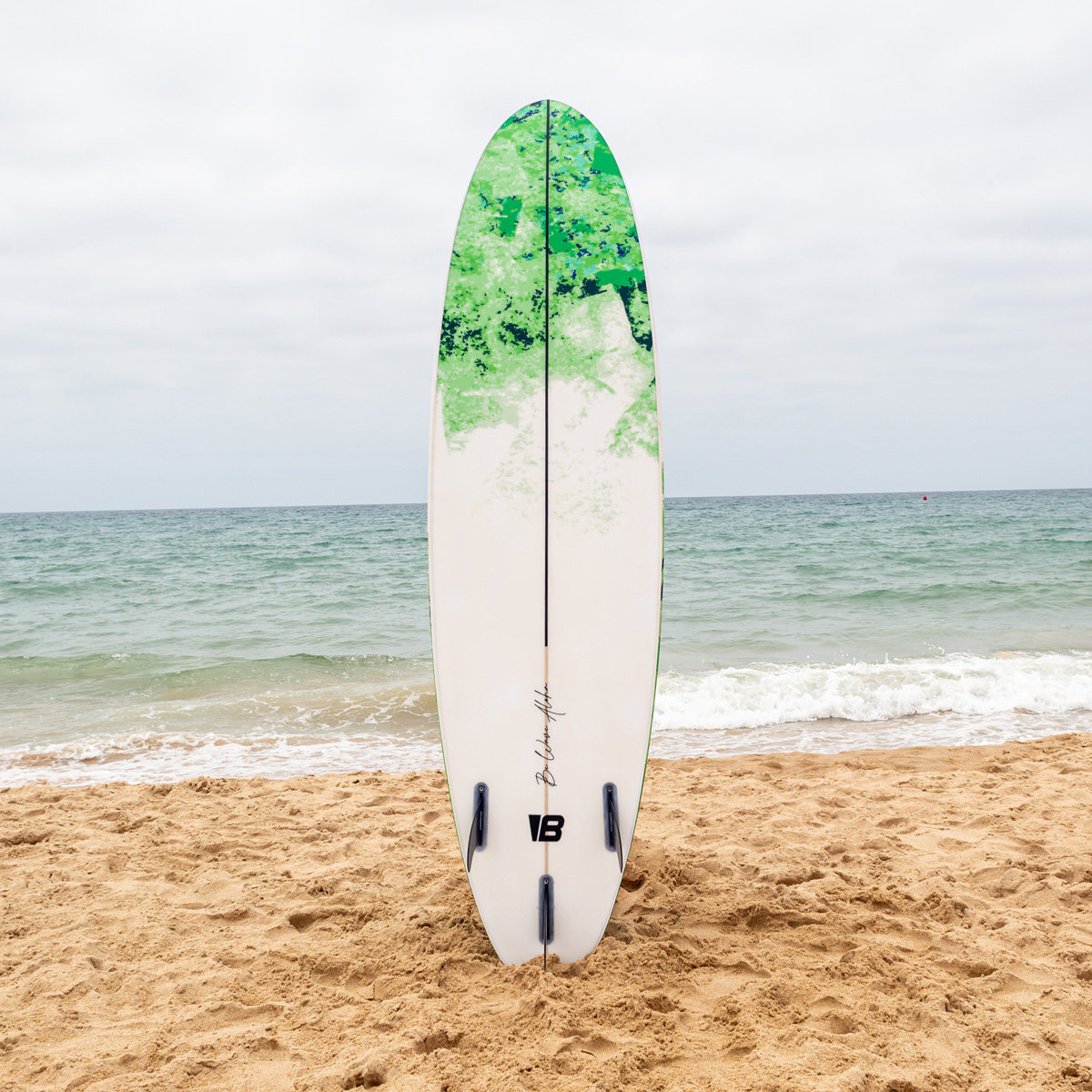 Una tabla de surf verde y blanca vertical en la arena de una playa con el mar y un cielo nublado al fondo.