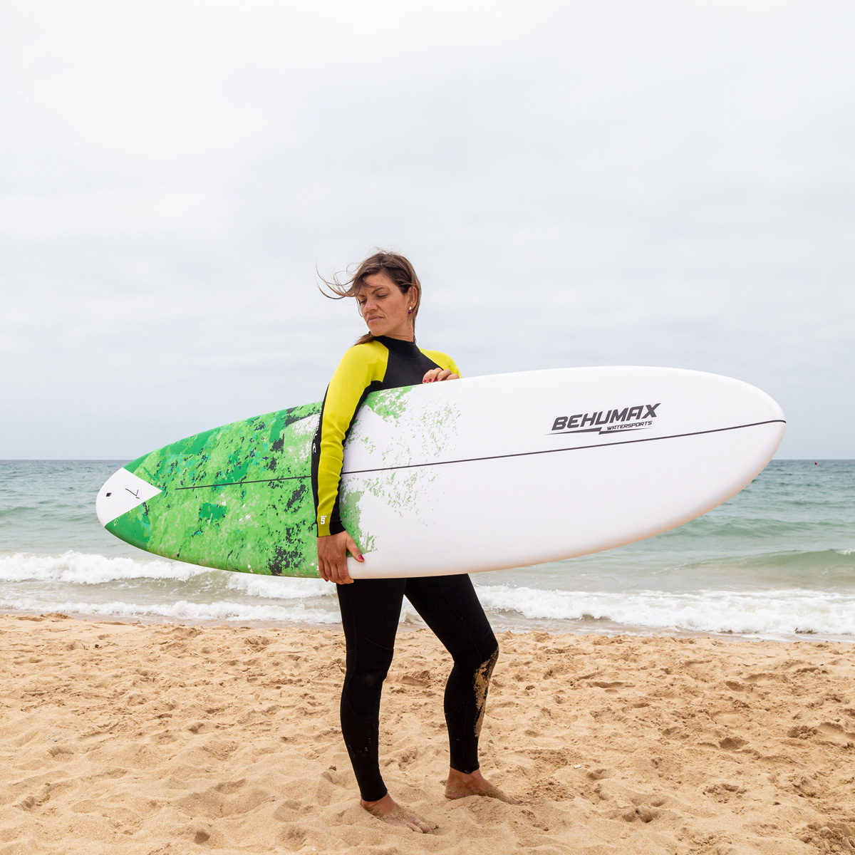 Una surfista con traje de neopreno negro y amarillo sostiene una tabla de surf blanca y verde en una playa de arena junto al mar.