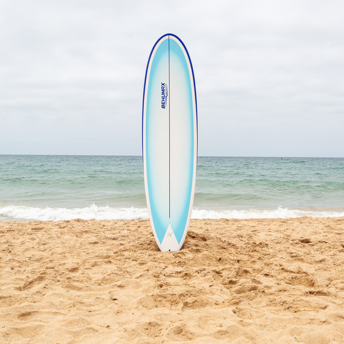 Una tabla de surf blanca y azul está clavada en la arena dorada de una playa con mar tranquilo y cielo nublado.