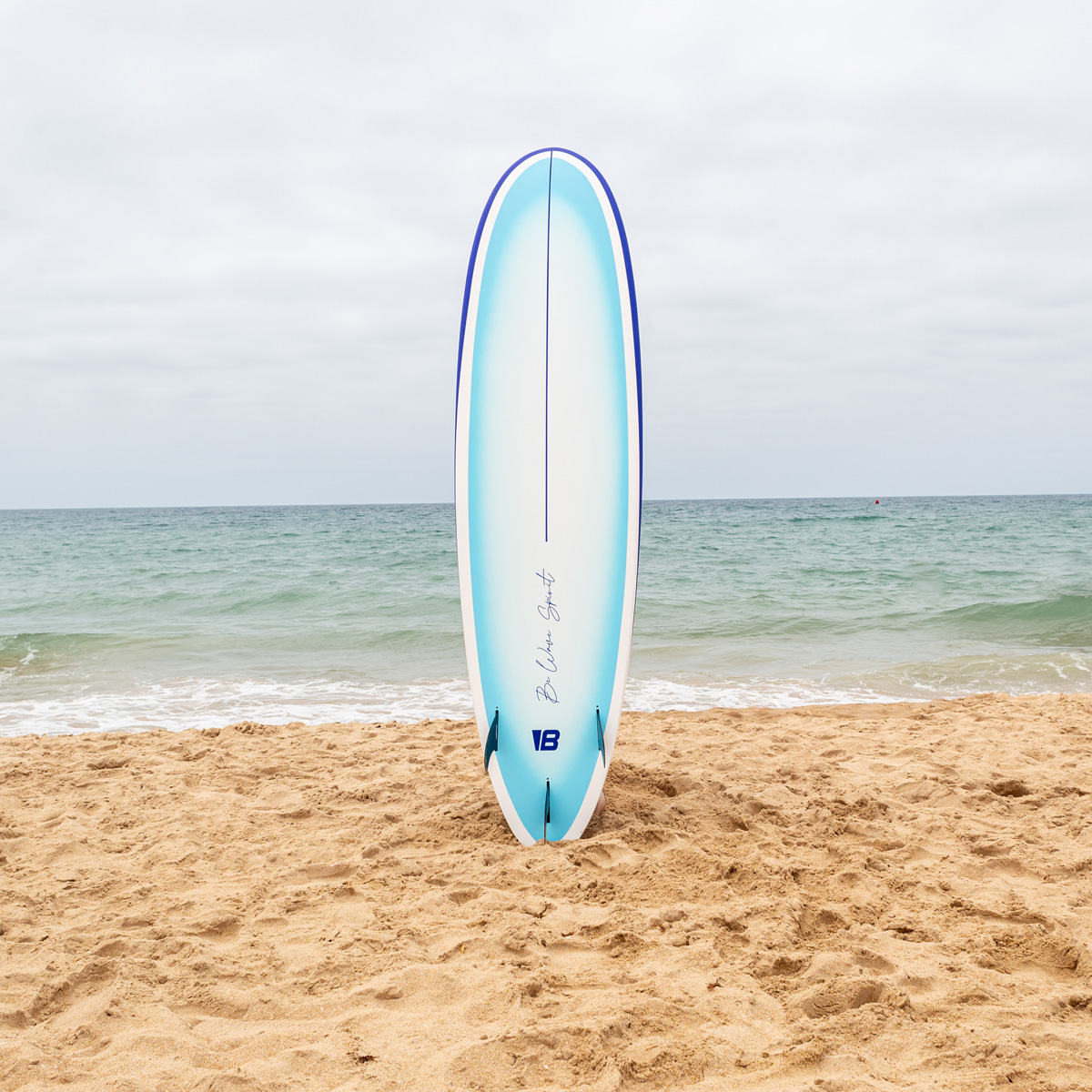 Una tabla de surf azul y blanca se encuentra verticalmente en la arena de una playa con el océano.