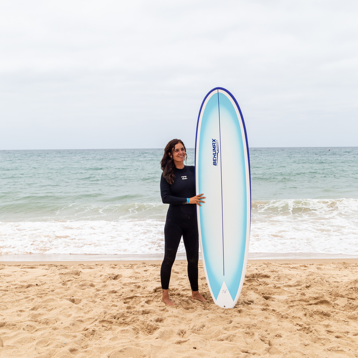 Una mujer sonriente en traje de neopreno negro sostiene una tabla de surf en una playa de arena con el mar de fondo.
