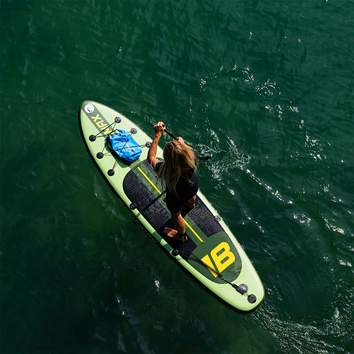Vista aérea de una mujer haciendo paddle surf en agua oscura y tranquila, con una bolsa azul.