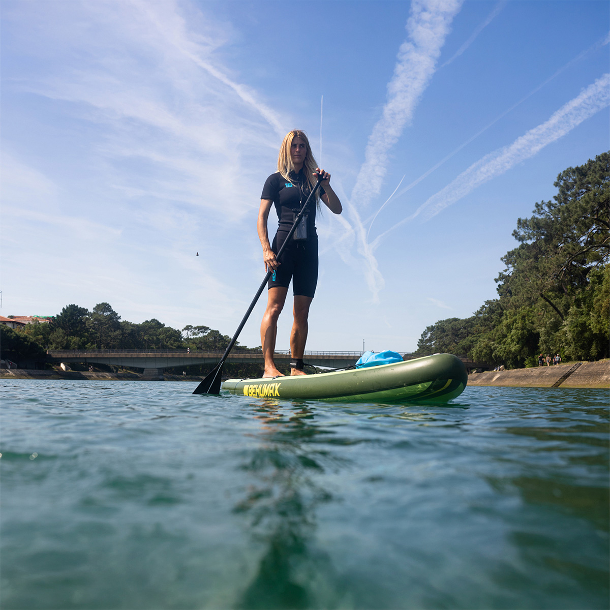 Una mujer de pie en una tabla de paddle surf verde sobre agua tranquila, con árboles al fondo y cielo azul.