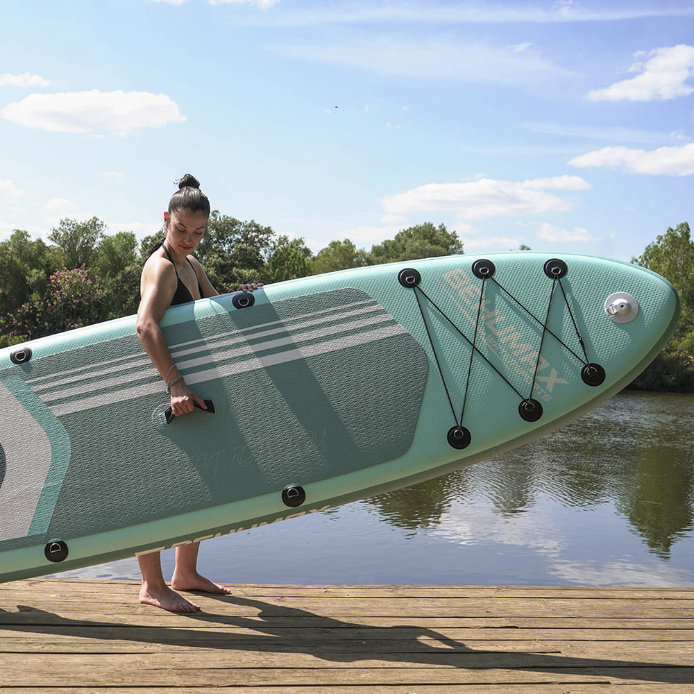 Una mujer lleva una tabla de paddle surf grande, de color turquesa y gris, en un muelle de madera junto a un lago.