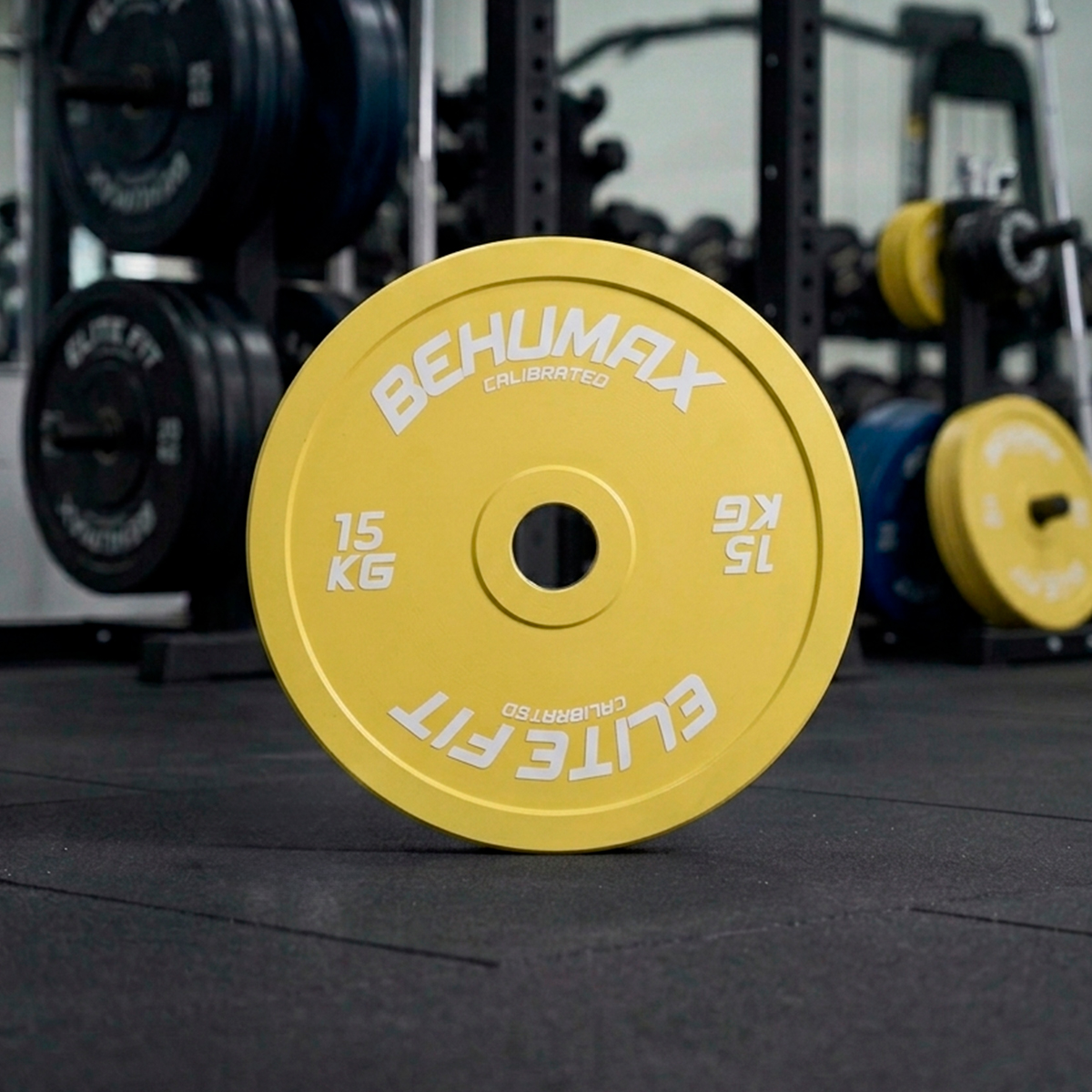 Un disco de peso amarillo de 15 kg está de pie en el suelo del gimnasio con bastidores al fondo.