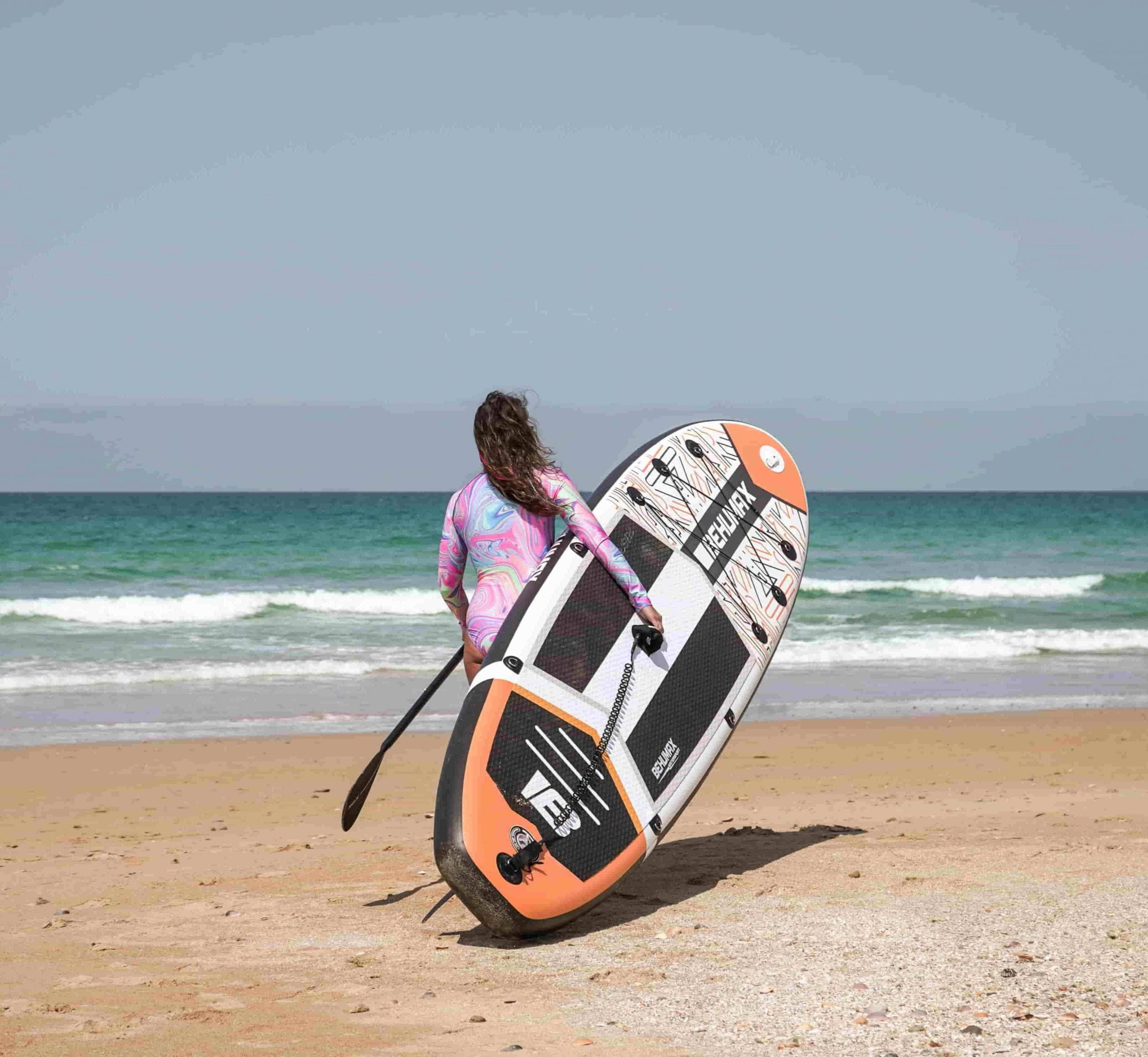 Una mujer en traje de baño colorido sostiene una tabla de surf de remo en la playa.
