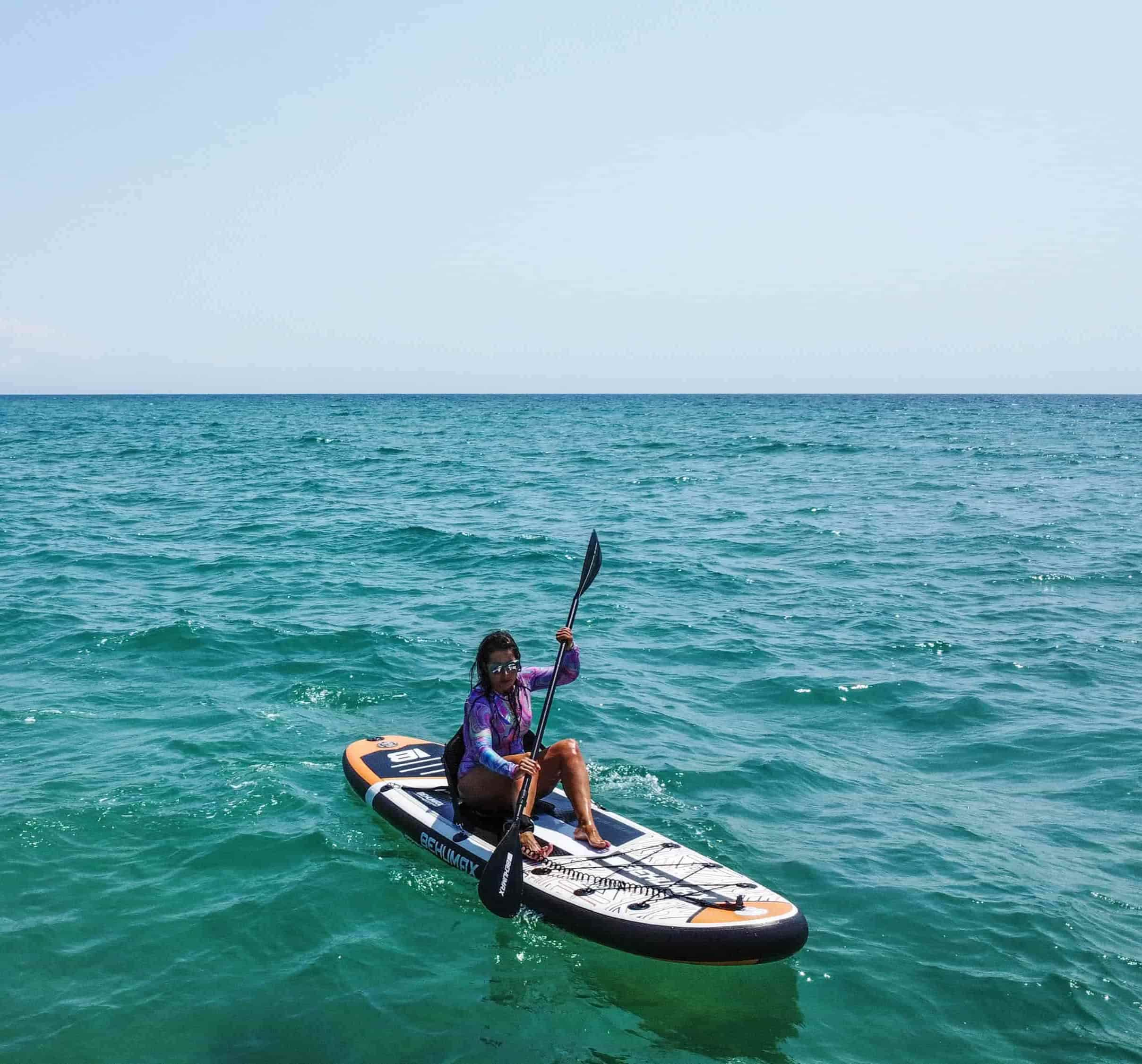Una mujer se sienta en una tabla de surf de remo, remando en el mar azul y tranquilo bajo un cielo despejado.