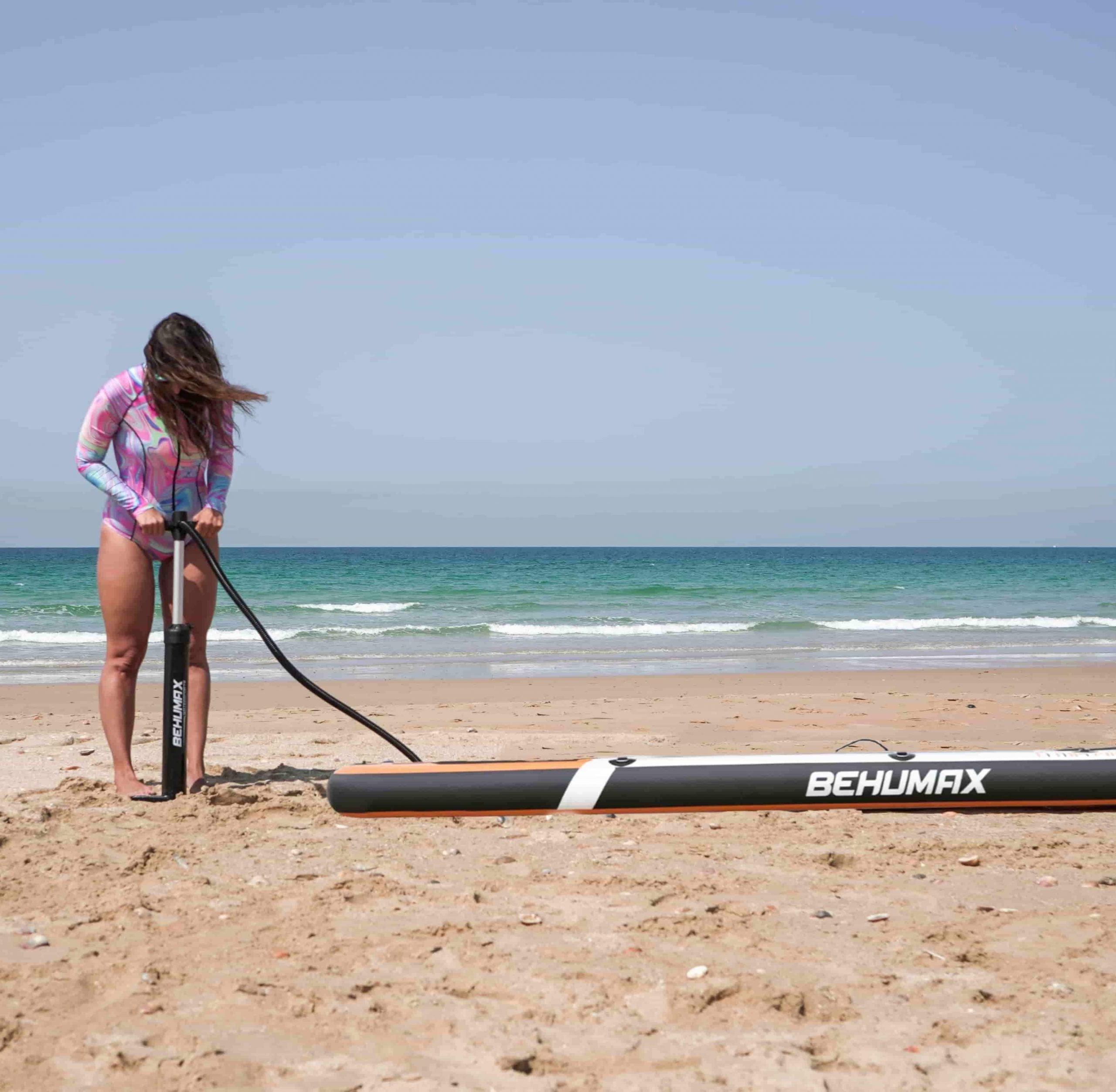 Una mujer en traje de baño colorido infla una tabla de SUP en la playa, con el mar azul de fondo.