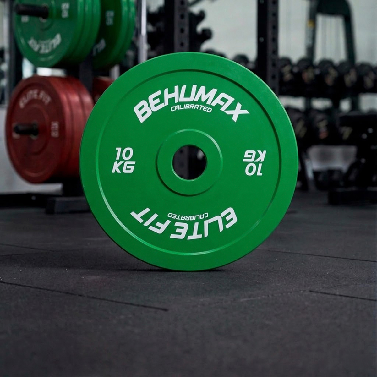 Un disco de pesas verde Behumax de 10 kg se sostiene verticalmente en el suelo del gimnasio con otros discos borrosos.