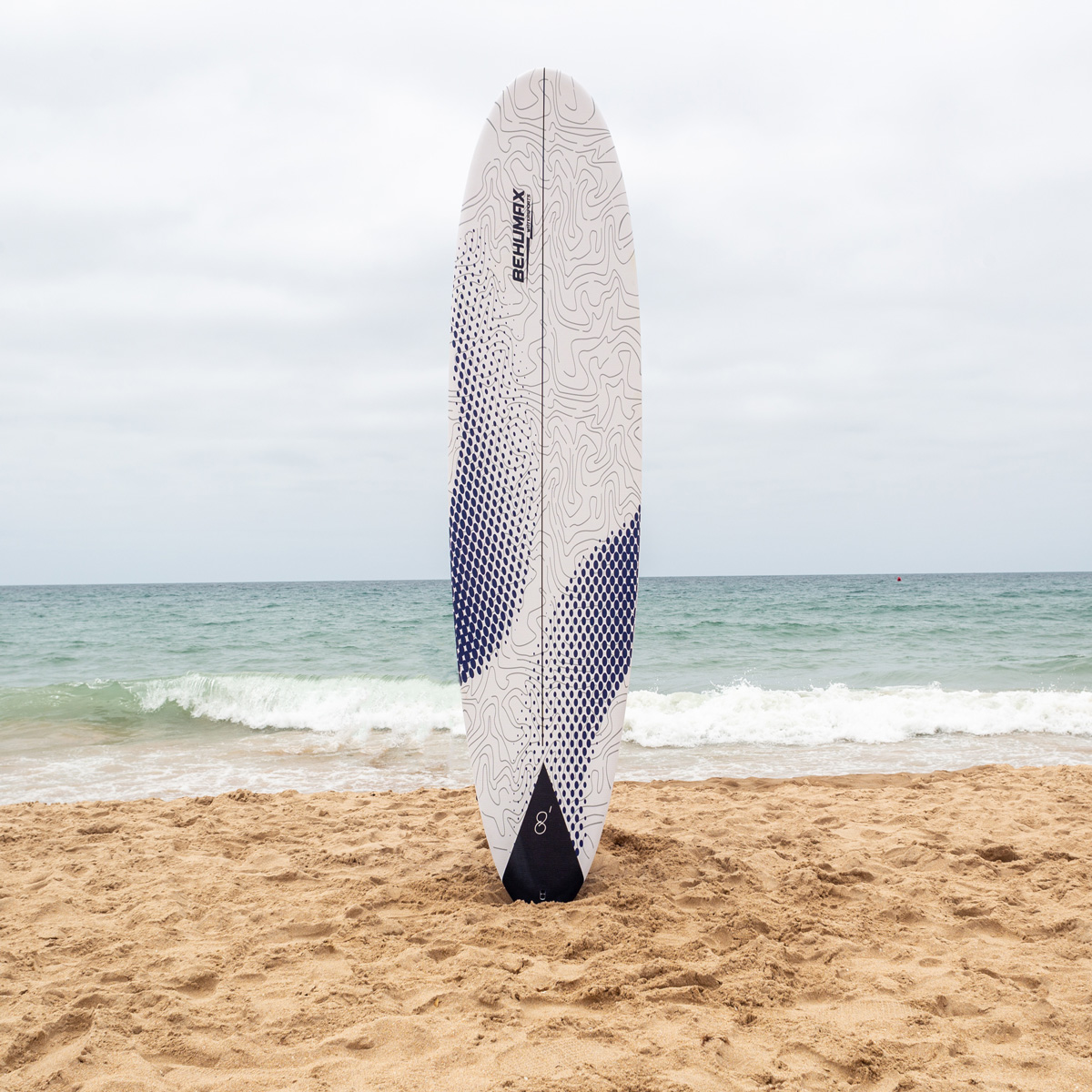 Una tabla de surf blanca con patrones de puntos azules se encuentra verticalmente en la arena de una playa con el océano al fondo.