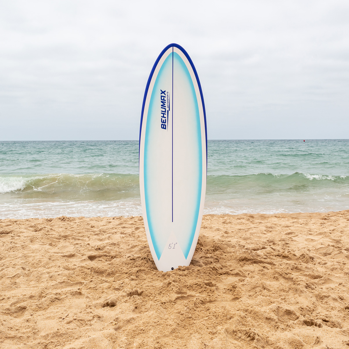 Una tabla de surf blanca con bordes azules y texto se encuentra en la arena de una playa, con el mar al fondo.