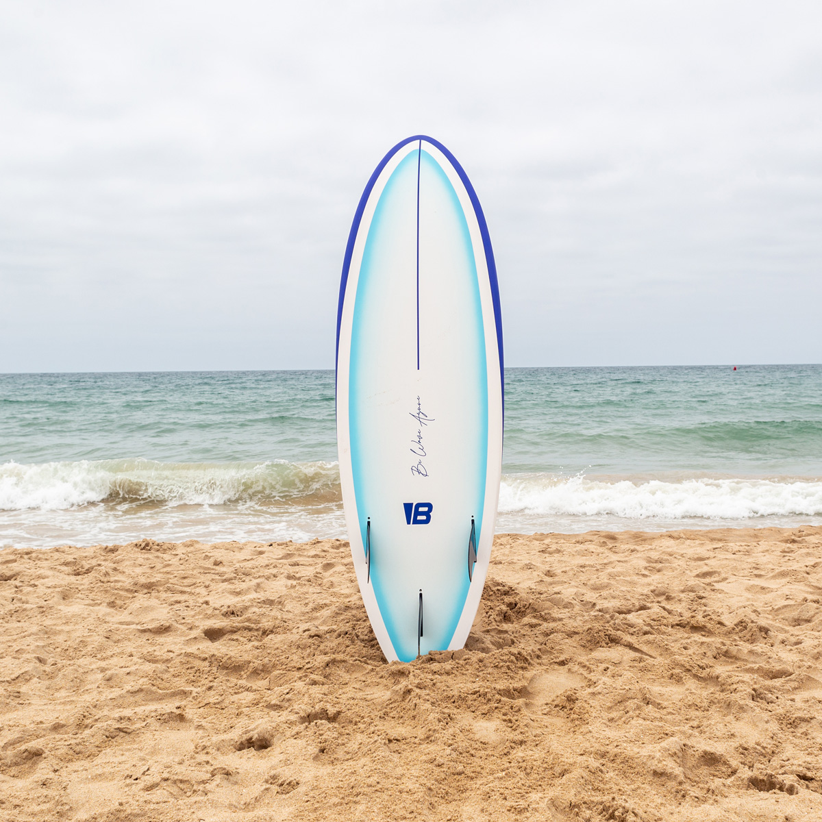 Una tabla de surf blanca y azul se encuentra verticalmente en la arena de una playa con el océano y el cielo nublado al fondo.
